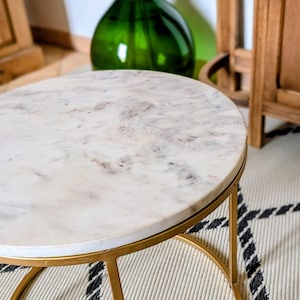 Op de afbeelding: Een ronde bijzettafel met een wit marmeren blad met grijze aders en een goudkleurig metalen frame. De tafel heeft een cirkelvormige voet en dunne, verticale metalen poten die het stenen oppervlak ondersteunen.