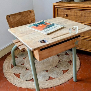 Peut inclure: Un bureau d'écolier vintage avec un plateau en bois clair, un compartiment de rangement intégré et des pieds en métal vert pâle. Le bureau présente un trou d'encrier circulaire, une rainure à crayon et une petite étoile sur le devant. Un livre GEOGRAPHIE est posé dessus.