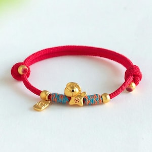 May include: Red cord bracelet with gold accents. The bracelet features a central charm with a bell and star design, along with small gold beads and a rectangular charm. The bracelet is a vibrant accessory.