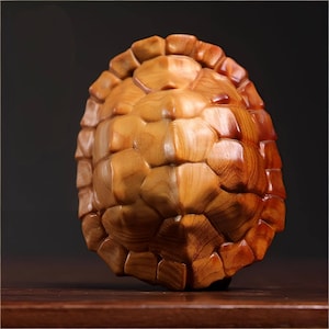 May include: A wooden sculpture of a turtle shell. The shell is carved from light brown wood, with a detailed, textured pattern. The sculpture has a smooth, polished finish, and the wood grain is visible. The overall shape is oval, and the sculpture is displayed on a wooden surface.