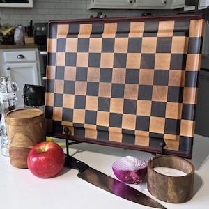 May include: A rectangular wooden chopping board with a dark and light brown checkerboard pattern, displayed on a black metal stand. The board is accompanied by a red apple, a silver chef's knife, a halved red onion, and two small wooden bowls.