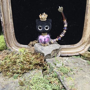 May include: A black cat figurine with large, round eyes and a gold crown. The cat has a pink and purple fluffy collar and is perched on a stone. A beaded tail with a heart charm curves around the cat.