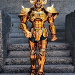 Può includere: Action figure dorata di un guerriero in armatura ornata, con un elmo con corna e punte sulle spalle. La figura è in posa su gradini di pietra, mettendo in risalto gli intricati dettagli dell'armatura.