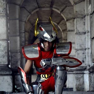 May include: A person in a red and silver cosplay costume, kneeling in a stone archway. The costume features a silver helmet with gold accents, red armor, and silver leg and arm guards. The overall aesthetic is futuristic.