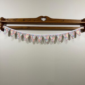 May include: A decorative bunting banner featuring triangular pennants with a repeating pattern of small, multi-coloured dots. The pennants are strung on a white ribbon and hang in front of a wooden shelf with a heart-shaped cutout.