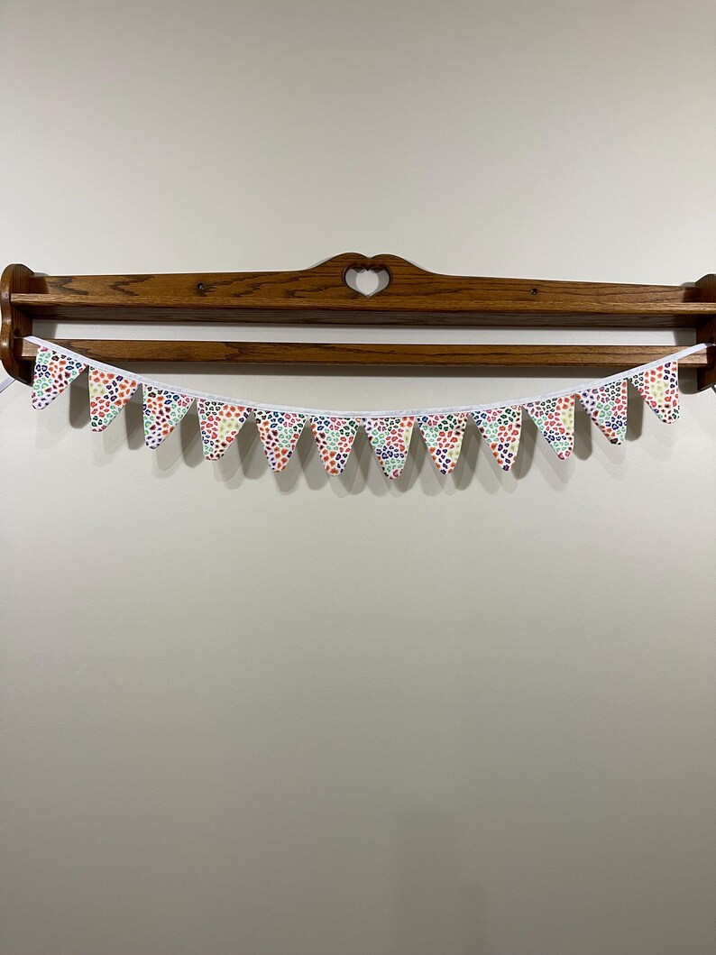 May include: A decorative bunting banner featuring triangular pennants with a repeating pattern of small, multi-coloured dots. The pennants are strung on a white ribbon and hang in front of a wooden shelf with a heart-shaped cutout.