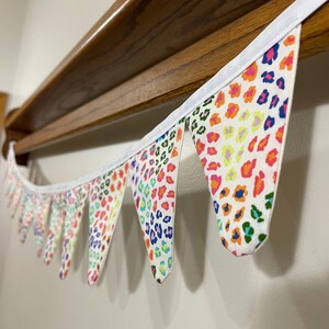 May include: A decorative fabric bunting with triangular flags. The flags display a leopard print pattern in pink, orange, yellow, blue, and green on a white background. The bunting is trimmed with white fabric.