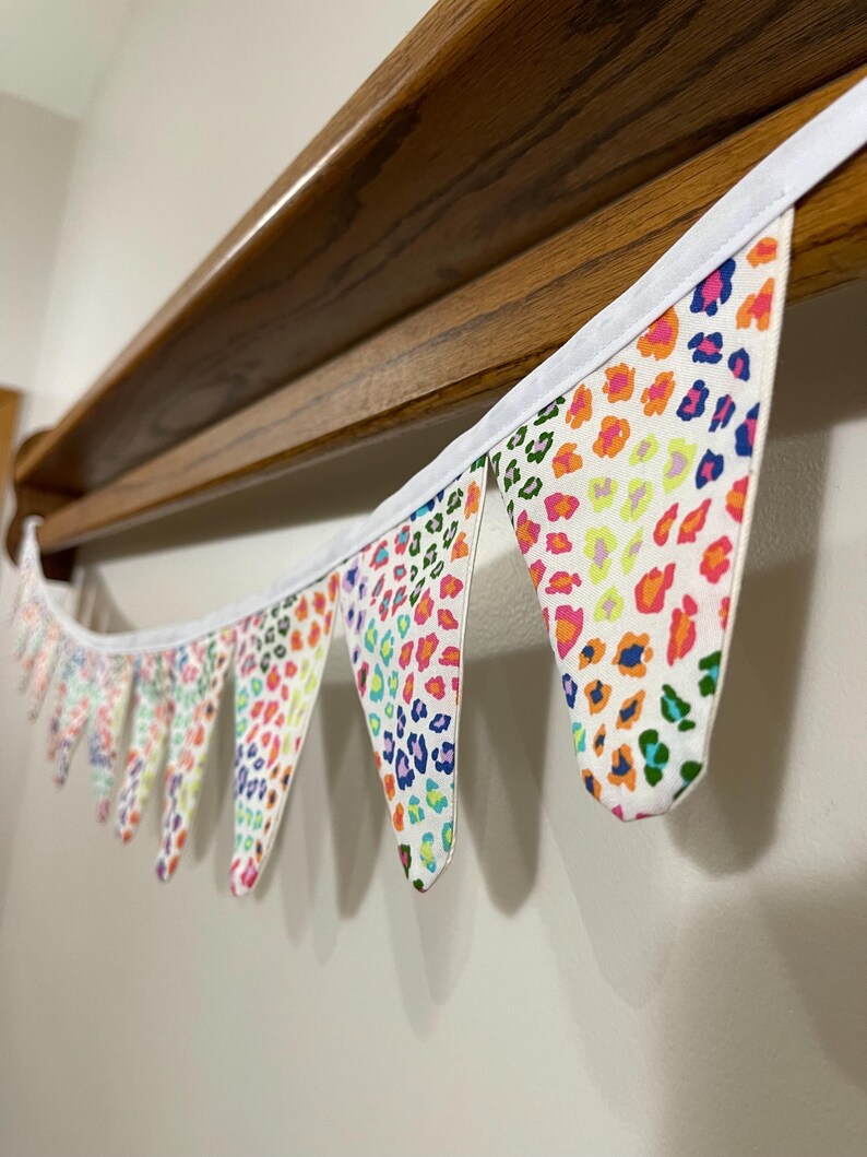 May include: A decorative fabric bunting with triangular flags. The flags display a leopard print pattern in pink, orange, yellow, blue, and green on a white background. The bunting is trimmed with white fabric.