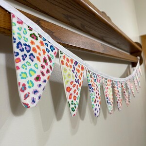 May include: A vibrant fabric bunting featuring a leopard print design in a range of colours including blue, pink, orange, and green. The bunting is strung on a white ribbon and hangs below a wooden shelf.