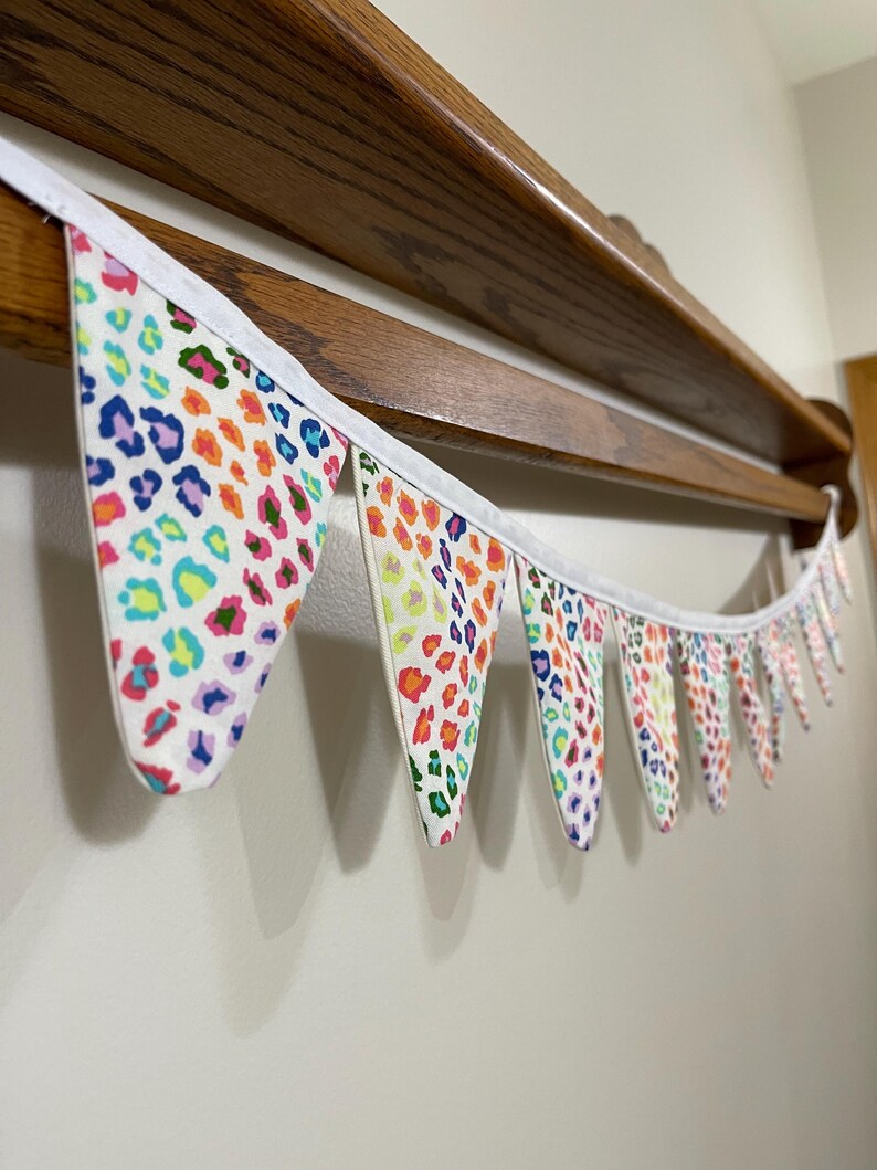 May include: A vibrant fabric bunting featuring a leopard print design in a range of colours including blue, pink, orange, and green. The bunting is strung on a white ribbon and hangs below a wooden shelf.