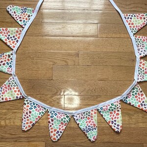 May include: A decorative fabric bunting with triangular flags. Each flag has a white base and a pattern of small, irregular shapes in red, orange, green, and blue. The flags are attached to a white ribbon.