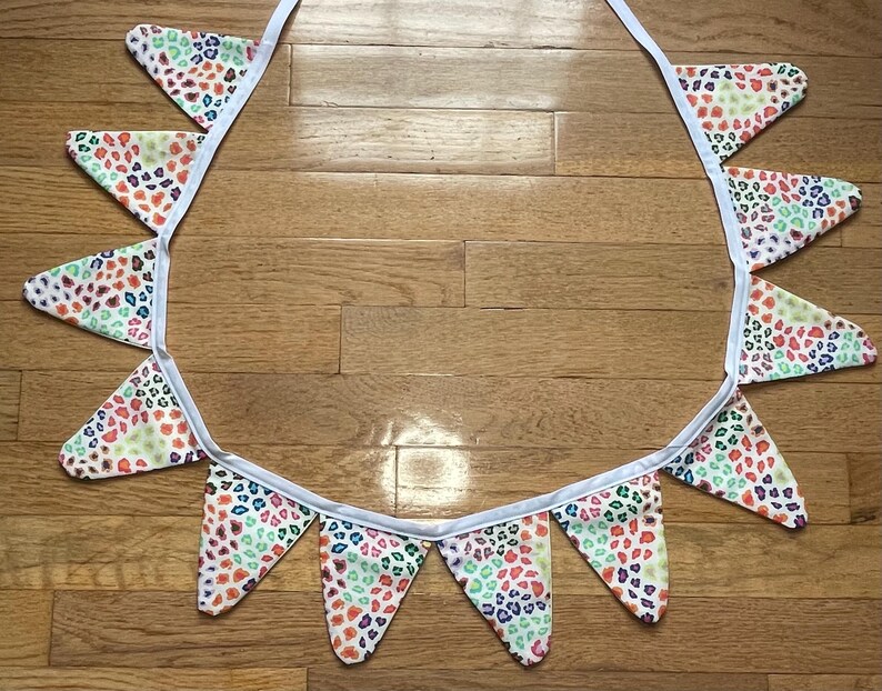 May include: A decorative fabric bunting with triangular flags. Each flag has a white base and a pattern of small, irregular shapes in red, orange, green, and blue. The flags are attached to a white ribbon.