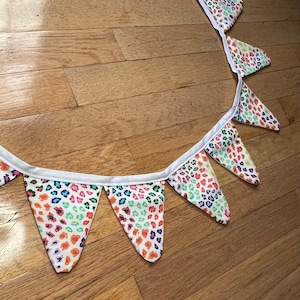 May include: A decorative fabric bunting with triangular flags. Each flag displays a vibrant leopard print pattern in orange, pink, blue, and green on a white base. The flags are joined by a white ribbon.