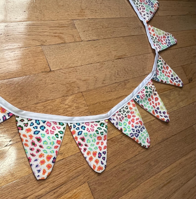 May include: A decorative fabric bunting with triangular flags. Each flag displays a vibrant leopard print pattern in orange, pink, blue, and green on a white base. The flags are joined by a white ribbon.