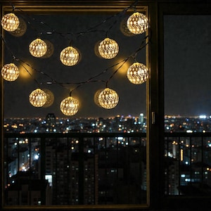 May include: String of decorative globe lights with intricate cut-out patterns, emitting a warm, inviting glow. The lights are strung across a window, with a cityscape visible in the background. The lights are a warm yellow color.