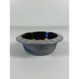 Puede incluir: Un cuenco de gres gris con interior oscuro y borde festoneado. El cuenco tiene detalles pintados en azul y un emblema estampado. El cuenco probablemente se utiliza para servir o exhibir.