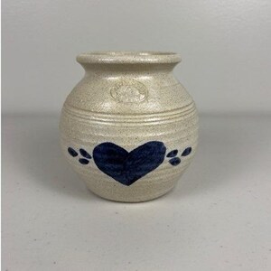 May include: A small, beige ceramic vase with a dark blue heart design. The vase has a rounded shape with horizontal lines and a small opening at the top. Decorative accents include dark blue leaf-like shapes.