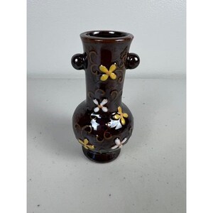 May include: A dark brown ceramic vase with a glossy finish, featuring two small side handles and a decorative pattern of raised yellow and white four-petalled flowers. The vase has a narrow neck, a bulbous body, and a flared rim at the top.