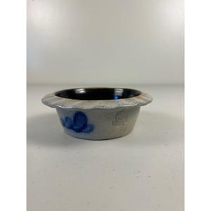 Puede incluir: Un cuenco pequeño y redondo de gres con borde festoneado. El cuenco es de color gris claro con interior oscuro y diseños florales azules. El cuenco tiene un sello en el lateral.