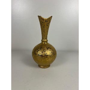 Könnte beinhalten: Eine goldfarbene Vase mit bauchigem Körper und schmalem, ausgestelltem Hals. Die Vase hat eine strukturierte Oberfläche mit dunkelbraunen Akzenten. Der Sockel ist rund und leicht erhöht. Die Vase ist wahrscheinlich aus Keramik oder Glas.