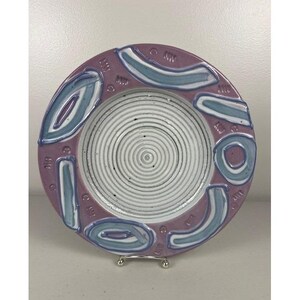 May include: A round ceramic plate featuring a white center with concentric circular grooves. The wide rim is a dusty mauve color, decorated with raised, abstract blue and white curved shapes and small circular indentations around the edge.