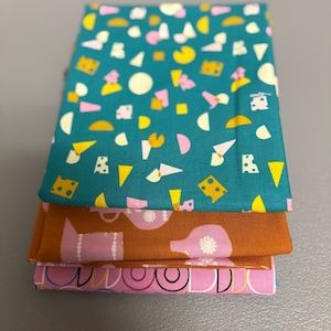 Peut inclure: Une pile de trois morceaux de tissu à motifs colorés. Le tissu supérieur est turquoise avec un motif répété de formes jaunes, roses et blanches. Le tissu du milieu est marron avec un motif de théière rose. Le tissu inférieur est rose avec un motif noir et blanc.