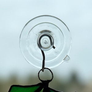 May include: A clear, circular suction cup with a metal hook attached. The suction cup is made of transparent plastic and has a small tab for easy removal. The hook is designed to hold a decorative item, such as a stained glass piece.