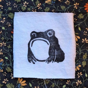 May include: A square, white fabric patch featuring a black frog linocut print. The frog has large eyes and a wide mouth. The background is a dark floral pattern.