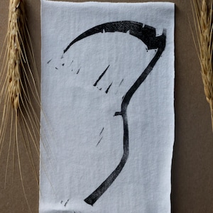 May include: A black scythe print on a white fabric square, set against a brown background. The image is framed with wheat stalks and lavender sprigs, creating a rustic, natural aesthetic. The scythe design is bold and graphic.