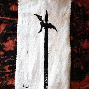 May include: A black illustration of a halberd printed on a white fabric square. The fabric has a raw, unfinished edge. The background is a mottled pattern of red and black.