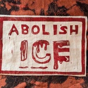 May include: A rectangular, off-white fabric with a red border and text that reads "ABOLISH ICE" in a bold, blocky font. The background is a mottled pattern of black and orange.
