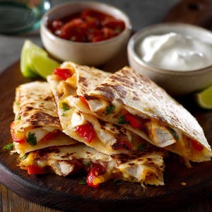 Puede incluir: Primer plano de quesadillas de pollo con pimientos rojos y queso, servidas en una tabla de madera. Dos cuencos pequeños de salsa y crema agria están en el fondo, junto con gajos de lima. Las quesadillas están cortadas en triángulos.