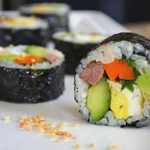 Op de afbeelding: Close-up van sushi rolls met rijst, zeewier en diverse vullingen, waaronder avocado, wortelen en vlees. De sushi is gerangschikt op een wit bord, met sesamzaadjes verspreid. De rollen zijn donker.