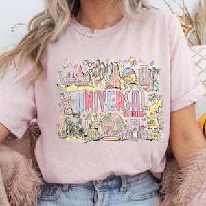 May include: A light pink t-shirt featuring a colorful graphic of Universal Studios attractions. The design includes the text "UNIVERSAL STUDIOS" in a multi-colored font, along with various cartoon illustrations.