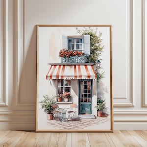 Könnte beinhalten: Aquarellmalerei eines europäischen Cafés mit einer rot-weiß gestreiften Markise, einem Balkon mit Blumen und einem kleinen Tisch und Stühlen. Das Gebäude hat eine grüne Tür und Fensterläden. Das Kunstwerk ist in einem hellbraunen Holzrahmen gerahmt.