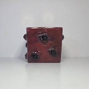 May include: A burgundy-colored, cube-shaped decorative object with three black, glossy, eye-like features on one side. The object has a textured surface and small, rounded protrusions on the corners.