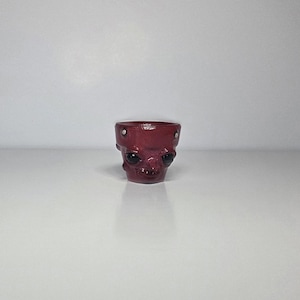 May include: A small, dark red, glossy ceramic planter shaped like a stylized alien head. The vessel features two large, smooth, black, oval-shaped eyes and two small, silver-colored circular studs embedded near the top rim.