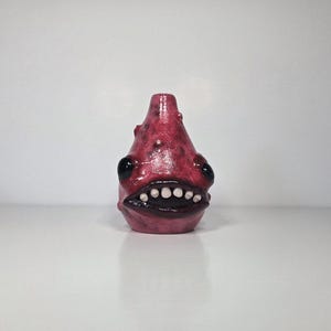 May include: A small, bulbous, reddish-pink ceramic sculpture shaped like a creature with a tapered top, two large black glossy eyes, and a wide mouth featuring dark lips and a row of small, rounded white teeth set against a smooth surface.