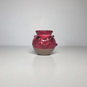 May include: A small, round ceramic vessel with a glossy red upper section and a matte beige base. The vessel features a sculpted face with two dark, protruding eyes and a wide, smiling mouth, along with small, rounded bumps on its sides.