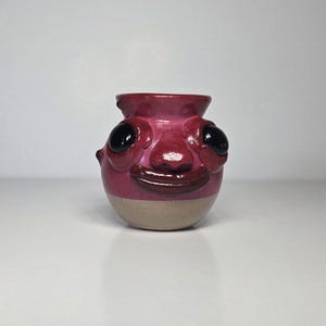 May include: A small, round ceramic vase featuring a sculpted face with large, glossy black eyes, a prominent nose, and a wide, dark red mouth. The upper portion is glazed in a vibrant magenta, while the base remains a natural, unglazed beige.
