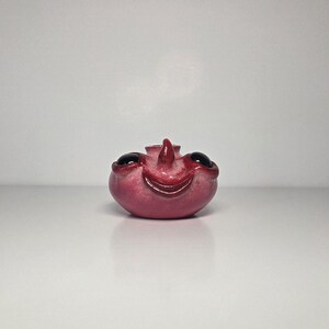 May include: A small, round, pink ceramic vessel featuring a sculpted face with a prominent nose, a wide smiling mouth, and two large, glossy black circular eyes set into the sides. The surface has a textured, mottled finish.
