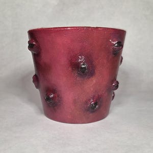 May include: A cylindrical, burgundy-colored ceramic pot with a textured surface. The pot features several dark, eye-like protrusions. The overall design gives the pot a unique, artistic appearance, suitable for decorative use.