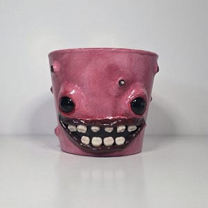 May include: A pink cylindrical ceramic planter featuring a wide, dark-colored mouth with visible white teeth, two large black glossy eyes, and small raised bumps across the surface. The vessel has a smooth, rounded rim and a flat base.