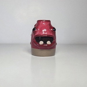 May include: A small, cylindrical ceramic vessel with a glossy magenta glaze on the upper portion and a matte beige base. The object features a wide, open mouth with two visible white teeth, and two dark, round eyes set into the surface.