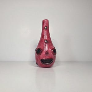 May include: A hand-sculpted ceramic vase in a deep magenta hue with a glossy finish. The vessel features a tapered neck, two small circular indentations, and a face with two dark, round eyes and a wide, open mouth with dark lips.