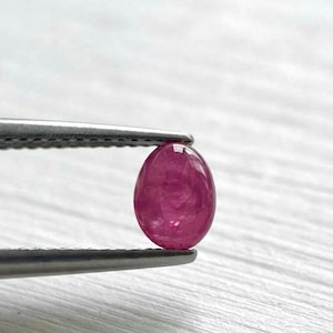Pode incluir: Uma pequena pedra preciosa de formato oval e cor vermelho-rosada vibrante, segurada por uma pinça de metal prateado. A pedra apresenta uma superfície lisa e polida com uma qualidade translúcida, refletindo a luz no seu topo arredondado.