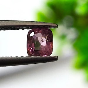 Puede incluir: Una gema de color rosa púrpura, talla cojín, es sostenida por unas pinzas. La gema, de forma cuadrada con esquinas redondeadas, refleja la luz. Las pinzas son de color gris oscuro. El fondo es borroso, verde y blanco.