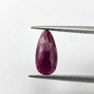 May include: A pear-shaped, faceted ruby gemstone with a deep magenta-red color held between the tips of a pair of silver-toned metal tweezers. The gemstone displays a geometric pattern of triangular facets across its surface.