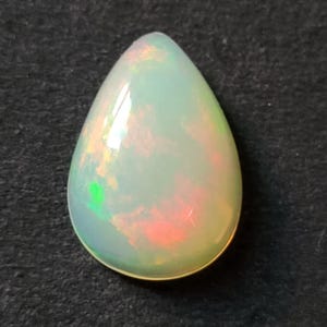 May include: A pear-shaped opal gemstone featuring a smooth, polished surface with a translucent white base color. The stone displays vibrant flashes of iridescent color, including bright green and warm orange-red hues throughout its interior.