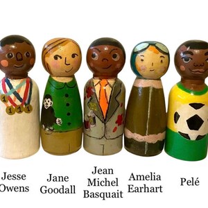 May include: Five hand-painted wooden peg dolls depicting Jesse Owens, Jane Goodall, Jean-Michel Basquiat, Amelia Earhart, and Pel&eacute;. Each doll is uniquely designed with detailed clothing and facial features. The names of each figure are written below them.